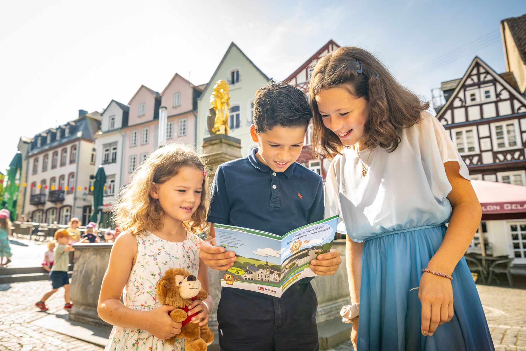 Drei Kinder stehen draußen auf einem sonnigen Stadtplatz mit bunten Gebäuden im Hintergrund und schauen sich gemeinsam eine Landkarte an. Ein Mädchen hält einen ausgestopften Löwen und lächelt, während sie die Gegend erkunden.